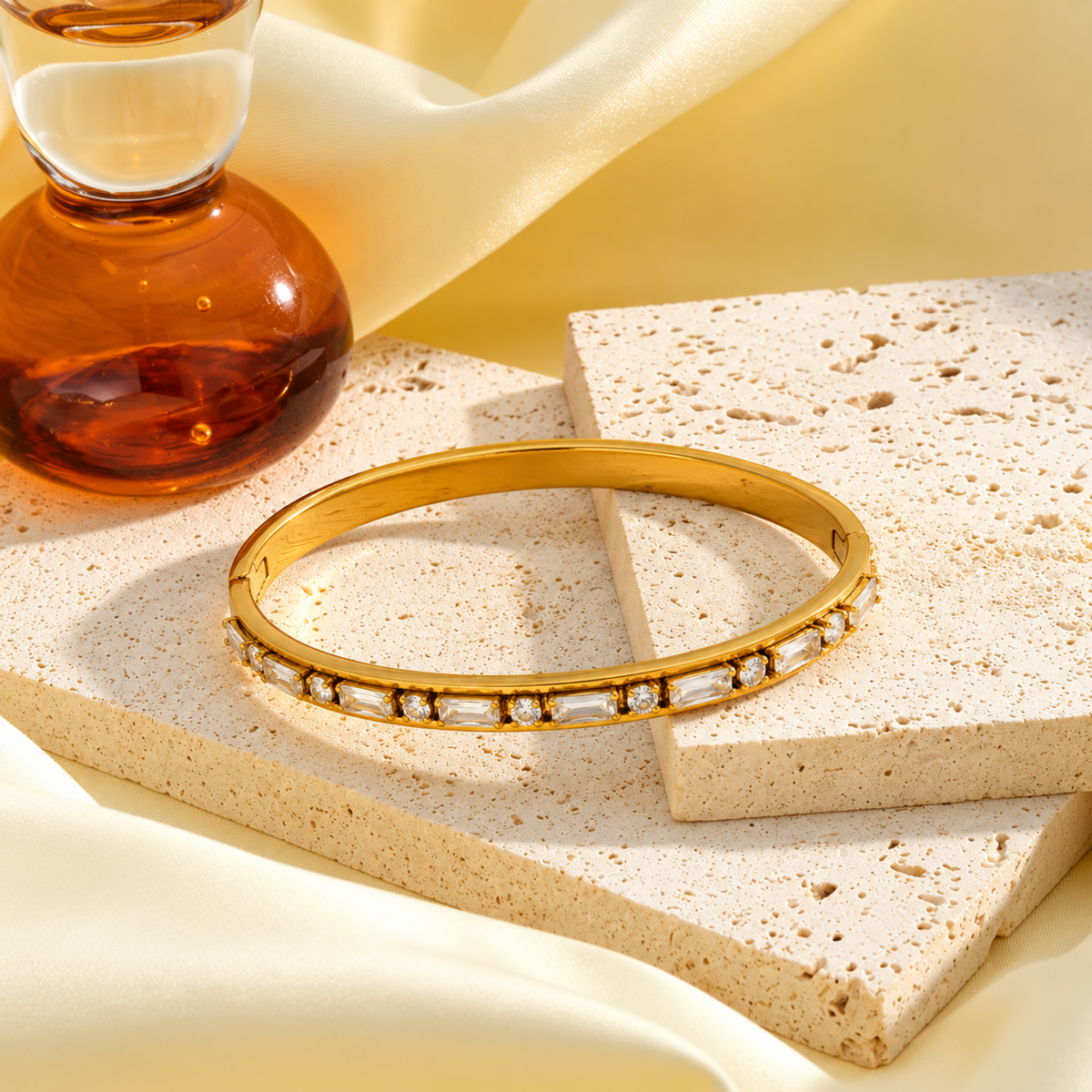 A gold bangle bracelet with a row of clear rectangular stones, displayed on stacked beige stone slabs with draped cream fabric and a glass bottle in the background.
