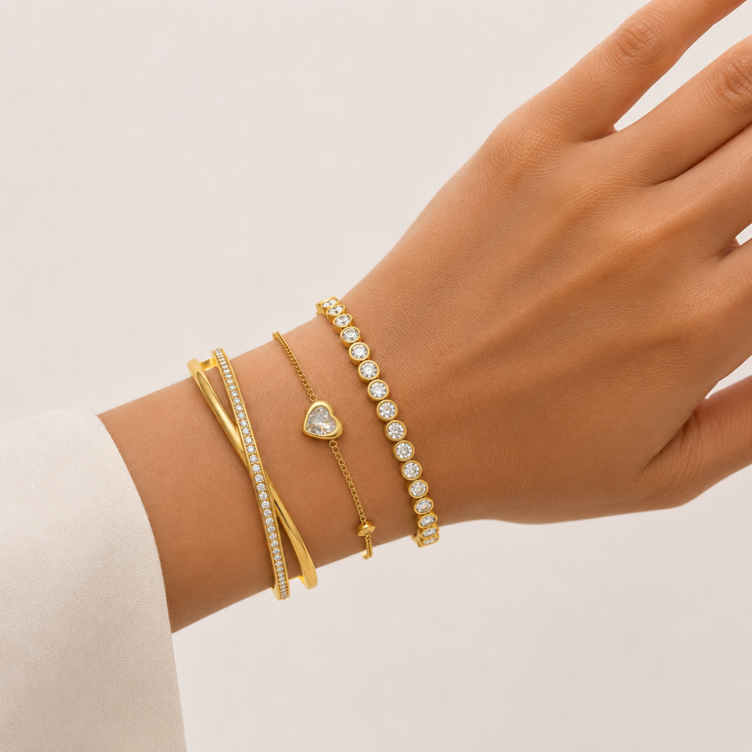 A premium three-piece gold-plated bracelet stack for women featuring a half-pavé crossover bangle, a sleek bezel-set crystal bangle, and a fine link chain with a single heart crystal charm, on a model