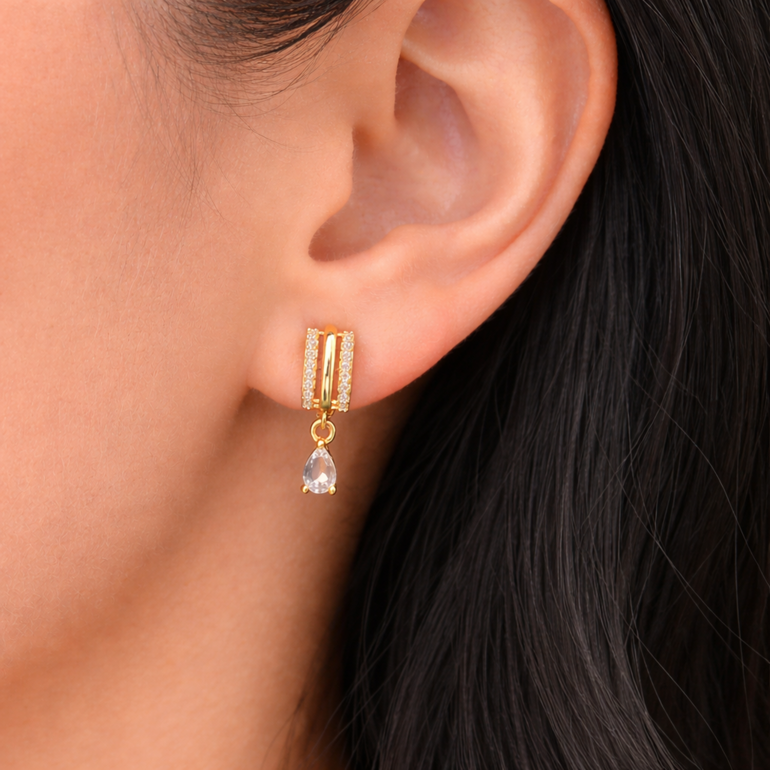 A gold-plated huggie hoop earrings for women featuring a suspended clear pear-shaped crystal drop, shown on a model