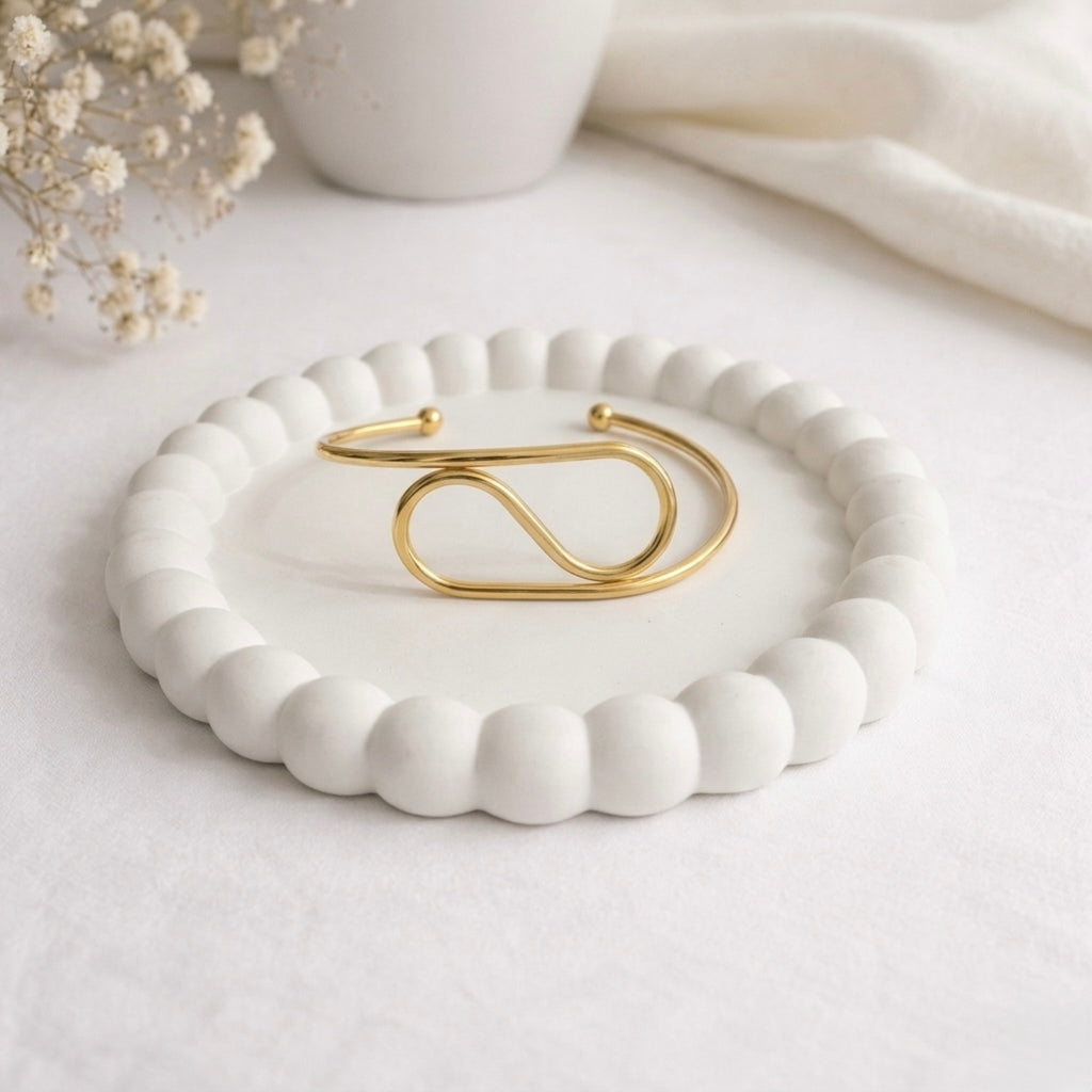 A modern gold-plated adjustable cuff bracelet for women featuring a sculptural infinite loop design, displayed on a white ceramic dish