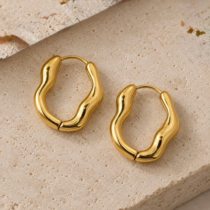 A pair of polished gold wavy hoop earrings displayed on a textured beige stone surface.