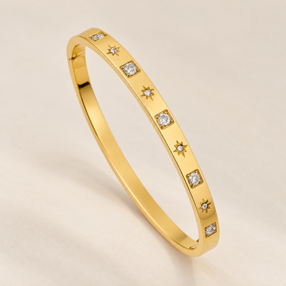 A yellow gold eternity-style bangle bracelet featuring small square-cut clear stones and starburst motifs set along the band, photographed on a clean light beige background.