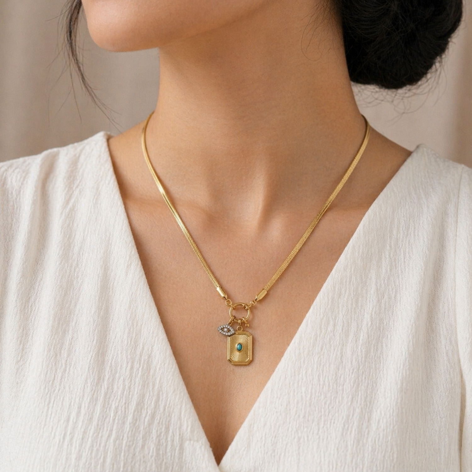 A gold-plated stainless steel chain for women featuring a flat snake chain and two protective charms, including an evil eye medallion, shown on a model