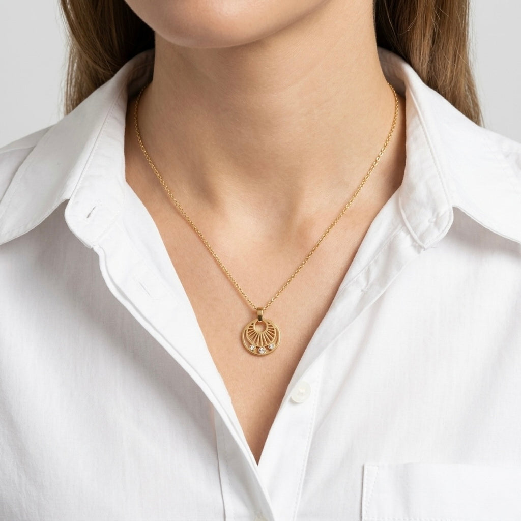 A gold-plated stainless steel necklace for women featuring a circular sunburst pendant with three small crystals at the bottom, shown on a model