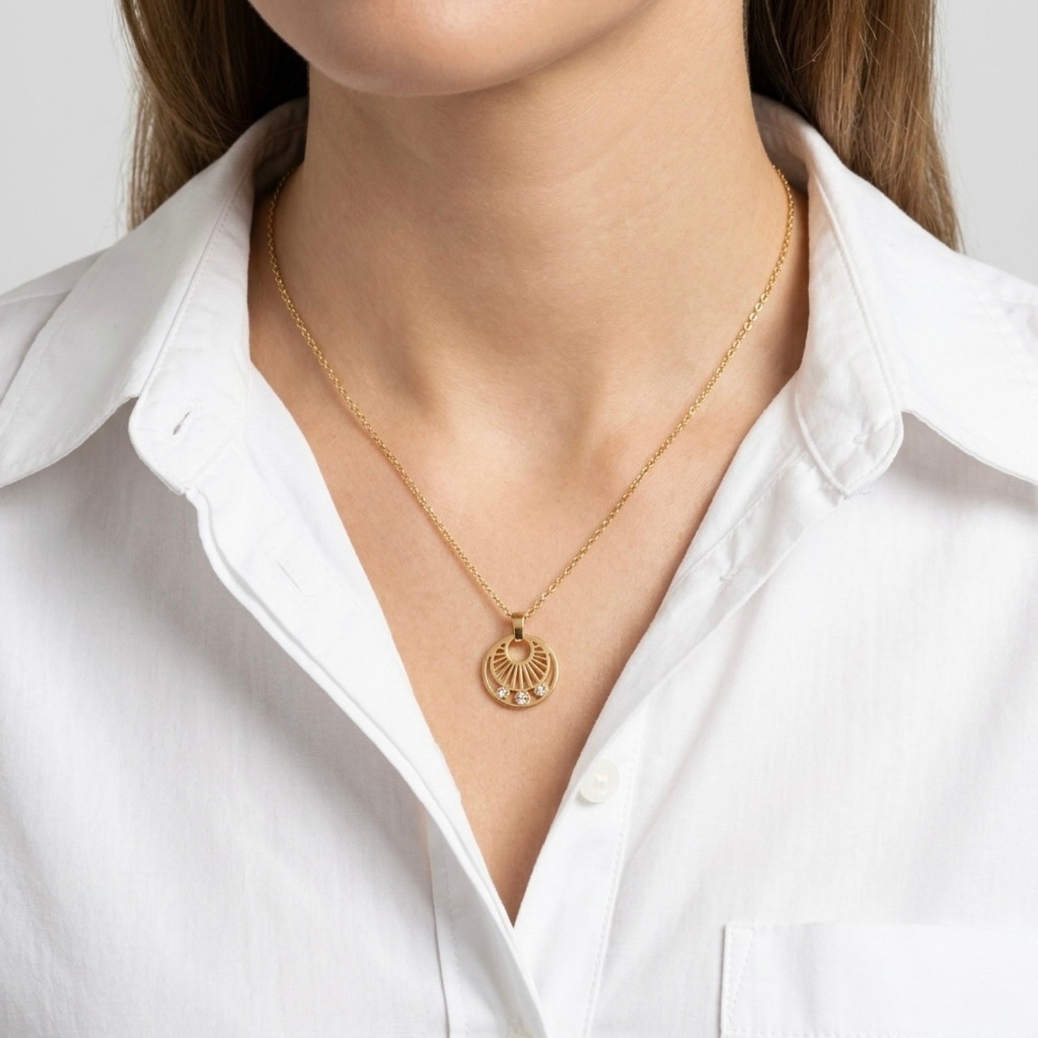 A gold-plated stainless steel necklace for women featuring a circular sunburst pendant with three small crystals at the bottom, shown on a model