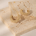 A pair of gold-plated hoop earrings for women featuring a prominent princess-cut clear crystal and smaller round accents on a textured background