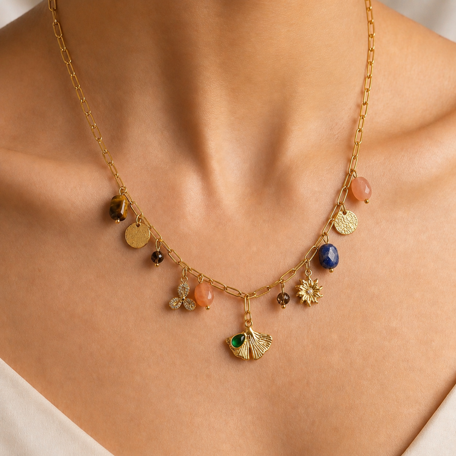 A gold-plated stainless steel chain for women featuring paperclip links and a variety of charms including a ginkgo leaf, lapis lazuli, and rose quartz beads, shown on a model