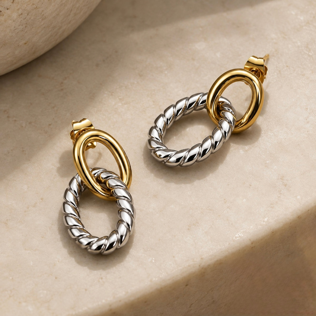 Two silver and gold drop earrings with twisted rope-style silver rings and smooth gold oval links, displayed on a beige stone surface.