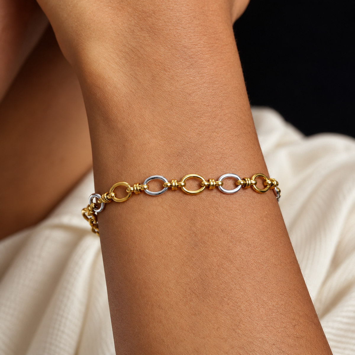 A close-up of a delicate two-tone chain bracelet worn on a person's wrist. The bracelet features alternating gold and silver oval links, shown against a soft cream-colored fabric and a dark background.