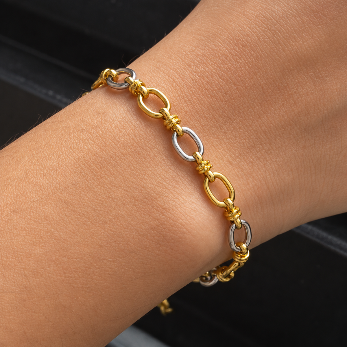 A close-up of a two-tone chain bracelet worn on a wrist. The bracelet features alternating polished gold and silver oval links connected by small twisted gold details.