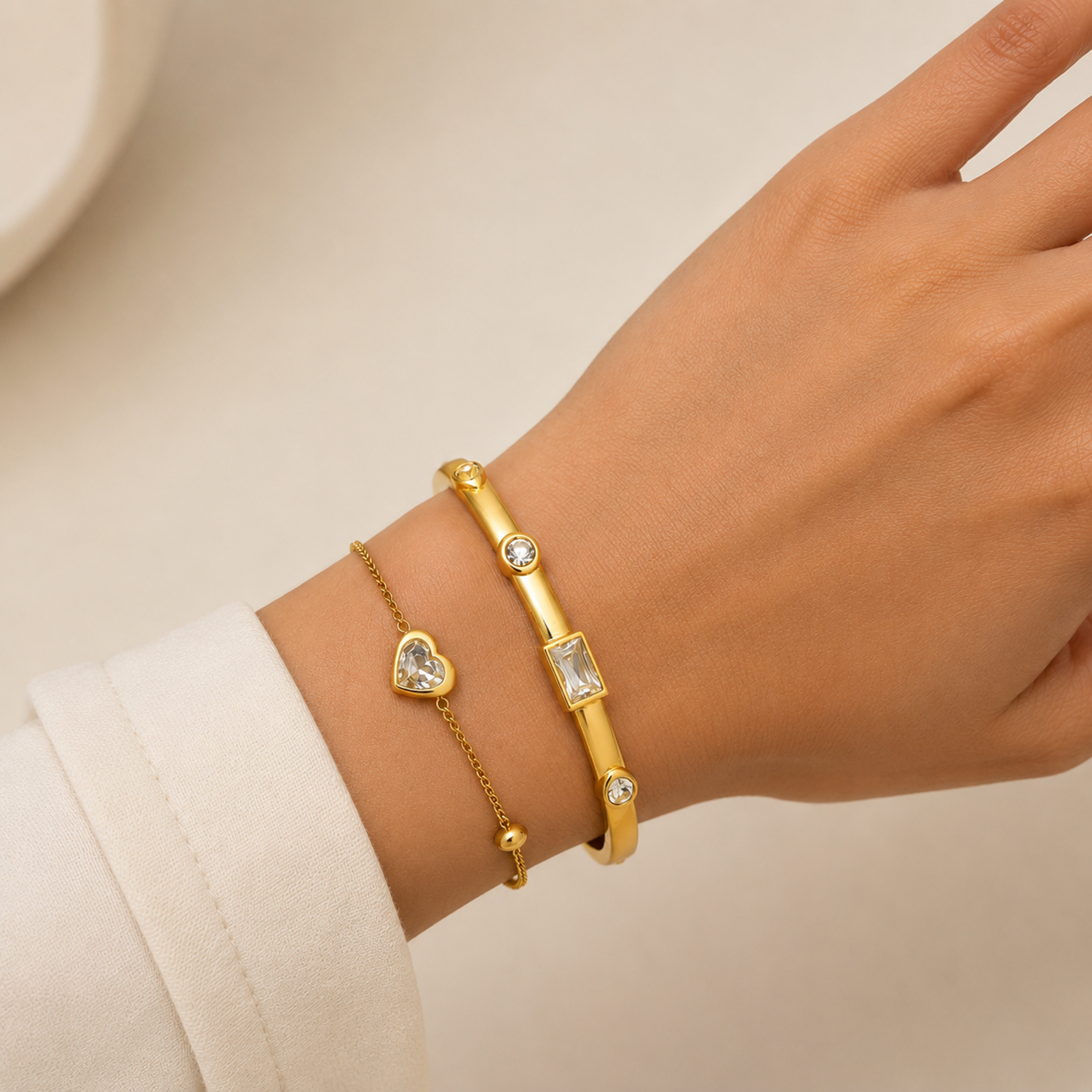 A premium two-piece gold-plated bracelet stack for women featuring a gold baguette station bangle and a fine link chain with a single heart crystal charm, on a model