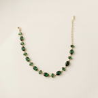 A gold-plated chain bracelet for women featuring a continuous row of deep emerald green oval-cut stones on a clean cream background