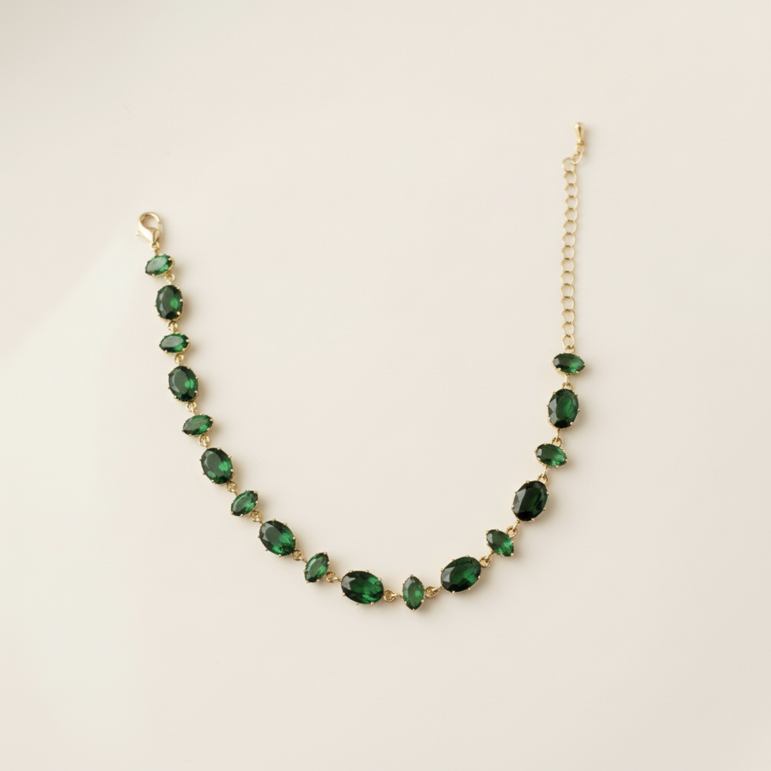 A gold-plated chain bracelet for women featuring a continuous row of deep emerald green oval-cut stones on a clean cream background
