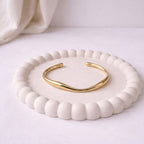 A minimalist gold-plated twisted cuff bracelet for women featuring wave design displayed on a textured white jewelry dish