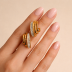 A pair of gold-plated triple-band hoop earrings for women featuring a central row of clear baguette crystals, shown on a model hand